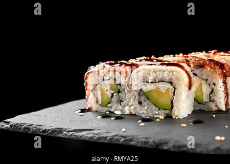 Close up of california roll filled with avocado and cream cheese, topped with unagi sauce. Sushi served on stone plate. Black background. Stock Photo