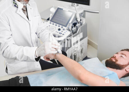 Bearded male patient lying in medical office, on ultrasound diagnostics. Professional therapist holding wrist of man, doing examination, using ultrasound probe and special equipment. Stock Photo