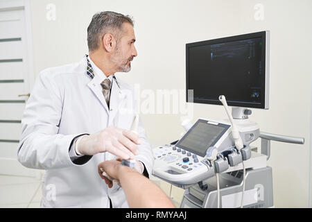 Mature and professional doctor holding hand of patient, doing ultrasound examination. Therapist using ultrasound probe for diagnosis. Physician looking at screen with results. Stock Photo