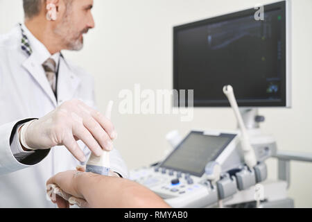 Mature doctor doing ultrasound diagnostics of patient's elbow joint. Therapist holding device and hand. Doctor wearing in white unifrom looking at screen, using ultrasound probe. Stock Photo