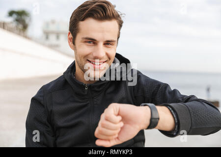 Image of joyful guy 30s in black sportswear looking at wrist watch while sitting on boardwalk at seaside Stock Photo