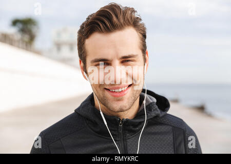 Image of sportive man 30s in black sportswear using earphones and mobile phone while walking along boardwalk at seaside Stock Photo