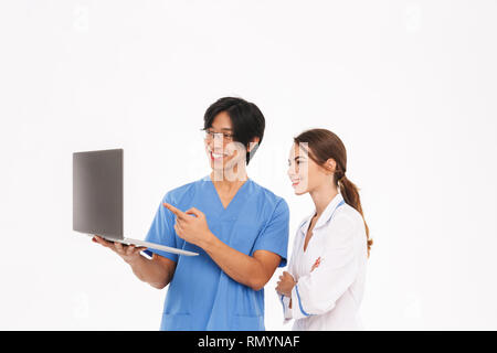 Smiling doctors couple wearing uniform standing isolated over white background, working on laptop computer Stock Photo