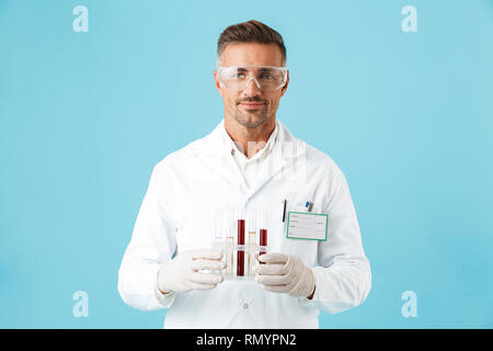 Confident man doctor wearing unifrom and glasses standing isolated over blue background, holding beakers Stock Photo