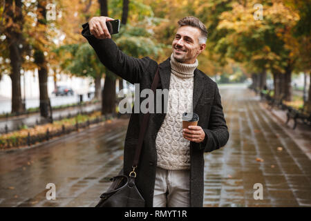Photo of joyful man 30s wearing warm clothes walking outdoor through autumn park and taking selfie on mobile phone Stock Photo