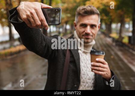 Photo of smiling man 30s wearing warm clothes walking outdoor through autumn park and taking selfie on mobile phone Stock Photo
