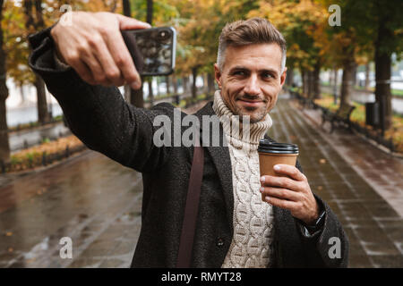 Photo of mature man 30s wearing warm clothes walking outdoor through autumn park and taking selfie on mobile phone Stock Photo
