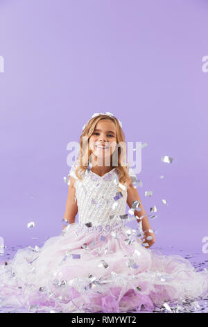 Pretty little girl dressed in princess dress sitting isolated over violet background Stock Photo