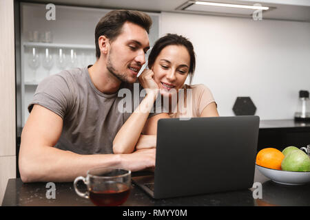 Photo of young couple man and woman sitting in kitchen and using laptop while having breakfast together Stock Photo