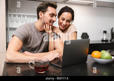 Photo of happy couple man and woman sitting in kitchen and using laptop while having breakfast together Stock Photo