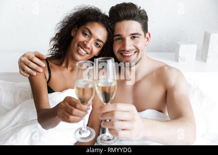 Beautiful happy young multiethnic couple relaxing in bed under blanket, drinking sparkling champagne from glasses Stock Photo