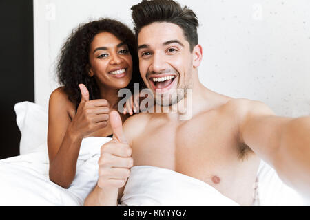 Beautiful happy young multiethnic couple relaxing in bed under blanket, taking a selfie Stock Photo