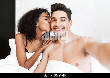 Beautiful happy young multiethnic couple relaxing in bed under blanket, taking a selfie Stock Photo