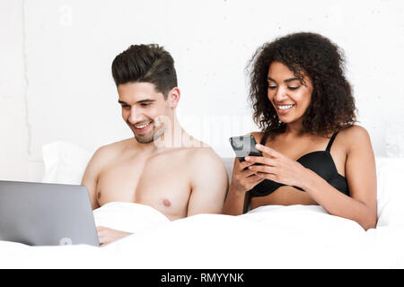 Beautiful happy young multiethnic couple relaxing in bed under blanket, man using laptop computer, woman holding mobile phone Stock Photo