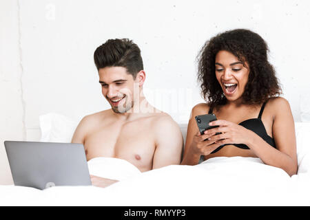 Beautiful happy young multiethnic couple relaxing in bed under blanket, man using laptop computer, woman holding mobile phone Stock Photo
