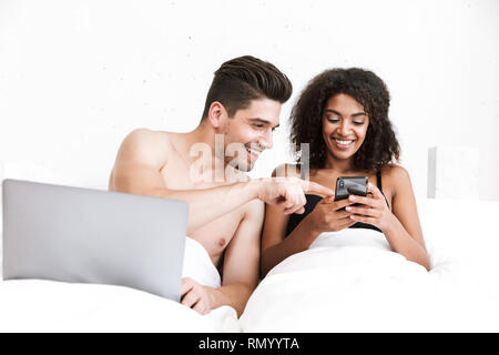Beautiful happy young multiethnic couple relaxing in bed under blanket, man using laptop computer, woman holding mobile phone Stock Photo