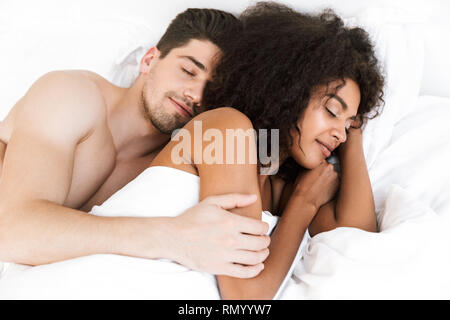 Beautiful happy young multiethnic couple relaxing in bed under blanket, woman sleeping Stock Photo