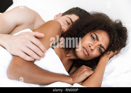 Beautiful young multiethnic couple relaxing in bed under blanket, woman frowning Stock Photo