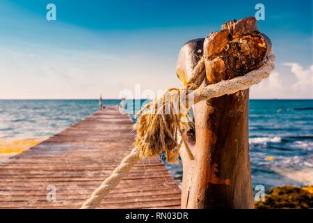 Old rustic wooden jetty out to the sea during a tropical sunny day on the caribbean coast of Mexico with old rope tied around post. Stock Photo