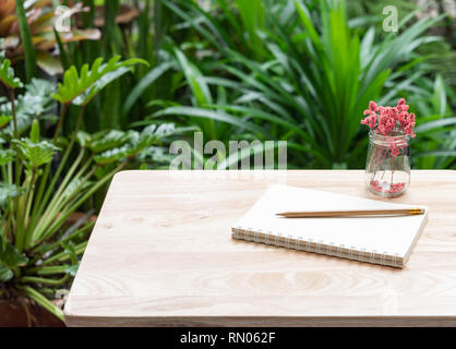 Notebook,brown pencil and beautiful red dried flower in glass vase on wood table with fresh green garden background,outdoor workspace Stock Photo