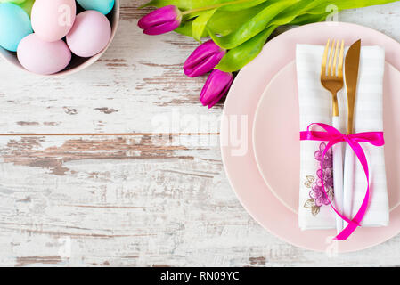 Easter table setting, flatlay with pastel colored Easter eggs, event decoration. Pink dishes and a gold fork and a spoon on a light rustic wooden back Stock Photo