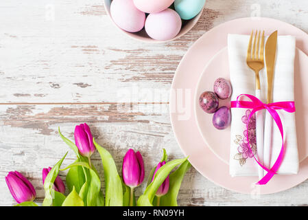 Easter table setting, flatlay with pastel colored Easter eggs, event decoration. Pink dishes and a gold fork and a spoon on a light rustic wooden back Stock Photo