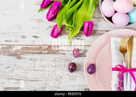Easter table setting, flatlay with pastel colored Easter eggs, event decoration. Pink dishes and a gold fork and a spoon on a light rustic wooden back Stock Photo