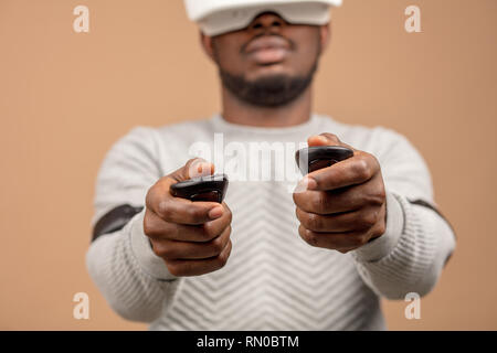 Dark-skinned millennial tourist in casual clothing wearing glasses with head-mounted display for smart phone, operating with controllers in hands, doi Stock Photo