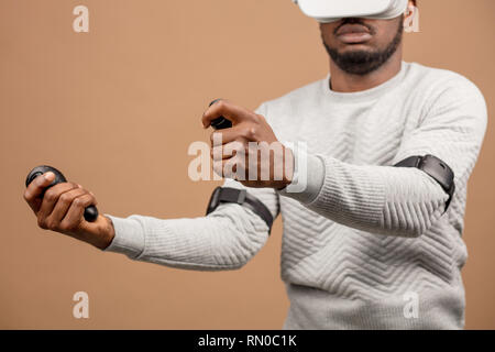 Dark-skinned millennial tourist in casual clothing wearing glasses with head-mounted display for smart phone, operating with controllers in hands, doi Stock Photo
