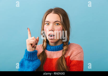 Shocked girl wearing sweater standing isolated over blue background, pointing up Stock Photo