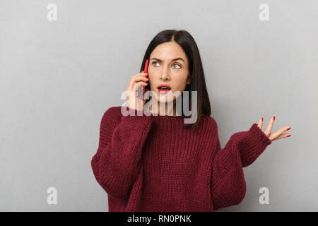 Picture of confused displeased young woman dressed in burgundy sweater talking by mobile phone isolated over grey wall background. Stock Photo