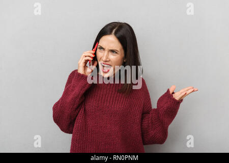 Picture of displeased young woman dressed in burgundy sweater talking by mobile phone isolated over grey wall background. Stock Photo