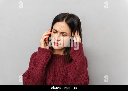 Picture of displeased young woman dressed in burgundy sweater talking by mobile phone isolated over grey wall background. Stock Photo
