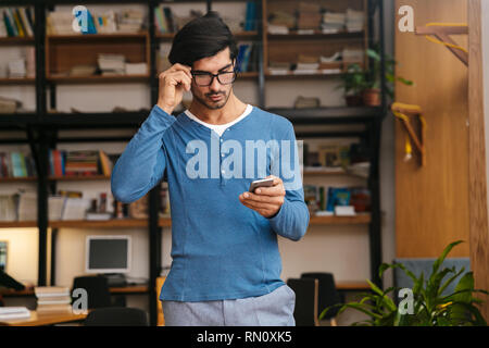 Serious handsome young man wearing glasses at the library, using mobile phone Stock Photo