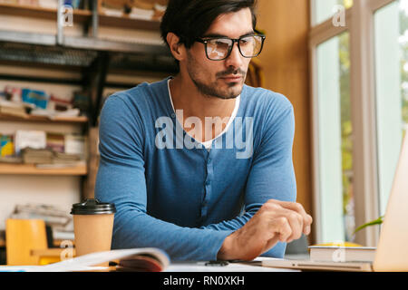 Handsome confident young man wearing glasses sitting at the library desk, working/studying, using laptop computer Stock Photo