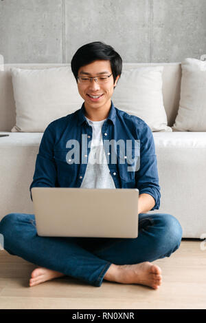 Happy asian man sitting at a couch at home, using laptop computer Stock Photo
