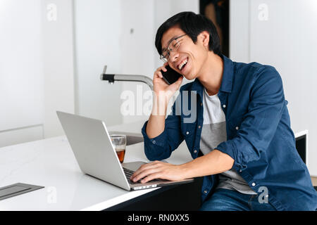 Smiling asian man working on laptop computer at home, talking on mobile phone Stock Photo