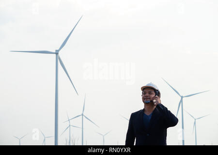 Successful asian engineer man standing and using smartphone with wind turbine. Stock Photo