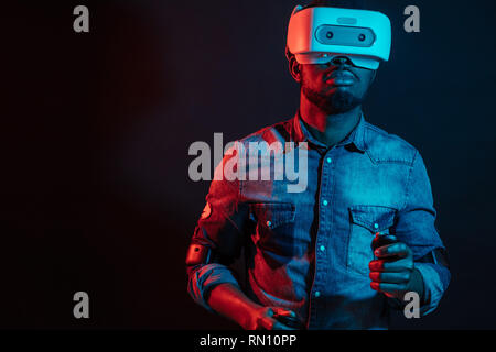 Unrecognizable young African man playing video game wearing virtual reality device against red and blue dual color light on dark background. Gaming eq Stock Photo
