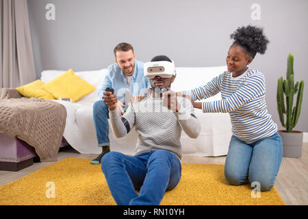 African male client testing 3D virtual reality headset playing video game while two diverse sales persons help him to take a purchase decision in shop Stock Photo