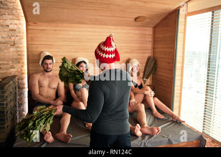Rear view of professional russian bath attendant in red hat, making show with two birch brooms before clients, preparing steam-room and people for ste Stock Photo