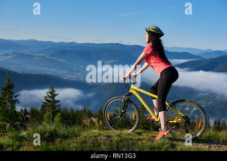 Happy young girl cyclist riding on yellow bicycle on a rural trail in the mountains, wearing helmet and red red t-shirt, enjoying morning haze in valley, forests on the blurred background. Copy space Stock Photo