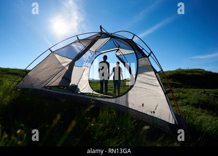 Back view of two young person resting near camping in the mountains under the sun on the blue sky. Guy and girl holding hands. View through a tent Stock Photo
