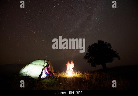 Night camping in mountains. Happy tourist girl sitting in entrance of lit from inside tent watching brightly burning campfire under amazing sky full of stars and Milky way. Tourism and travel concept Stock Photo