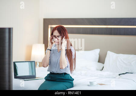 Tired woman with long ginger hair, wearing pencil skirt and blouse, sits on bed after working day and rubbing temples, feeling headache, or feeling st Stock Photo