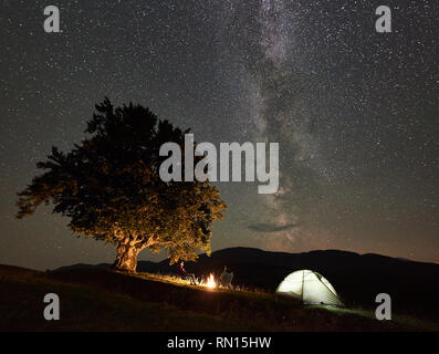 Young couple hikers enjoying night camping in mountains. Back view of man and woman sitting on chairs beside bonfire, illuminated tourist tent and big tree under starry sky full of stars and Milky way Stock Photo