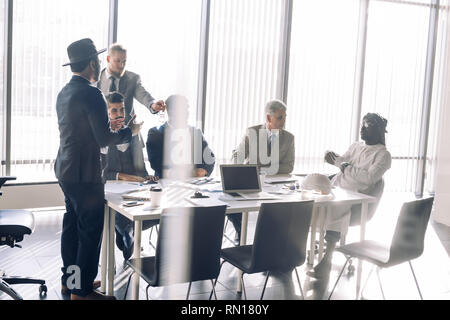 Multicultural business partners meeting discussing, using internet, and documents with graphs and charts, collaborating vively over panoramic windows  Stock Photo
