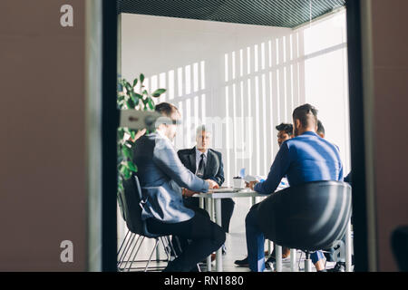 Mirror reflection shows multiracial male business executives discussing project sitting at conference table, men dressed in formal wear and have posit Stock Photo