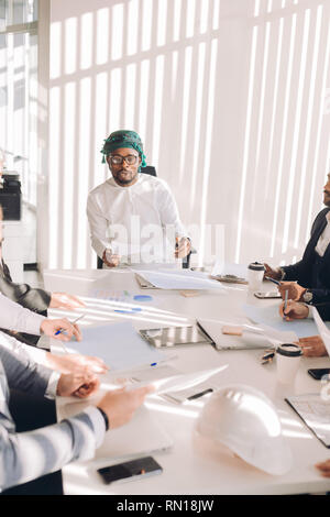 Multiracial group of experts headed by Arab leader, dressed in national white wear, analyzing business result and discussing profit. Stock Photo