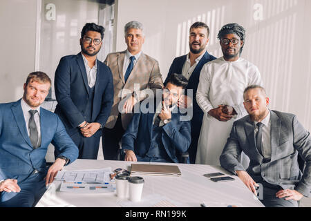 Handsome young bearded caucasian boss in a stylish suit and his european, indian and arabian partners, successful multiracial business team, taking se Stock Photo